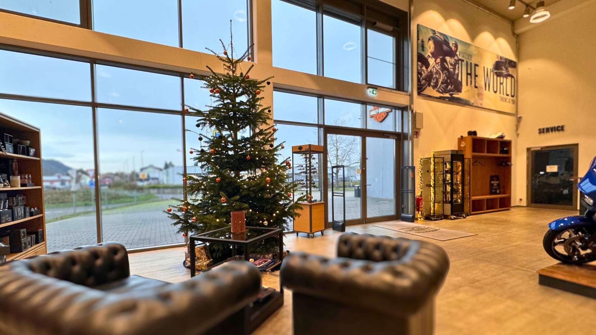 Ein geschmückter Weihnachtsbaum von innen fotografiert vor der gläsernen Ladenfront eine Harley-Davidson Händlers mit zwei Sesseln und Geschenken davor