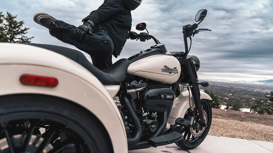 Ein Motorradfahrer sitzt auf einem weißen Harley-Davidson-Motorrad und hebt eine Bein auf die Sitzbank, während er die Aussicht auf eine bewaldete Landschaft genießt.