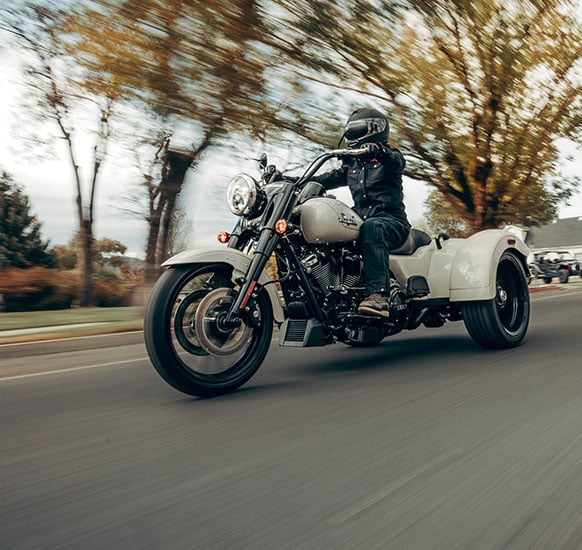 Ein Fahrer auf einem modernen Harley-Davidson Freewheeler fährt bei herbstlichem Wetter auf einer Straße, umgeben von Bäumen.