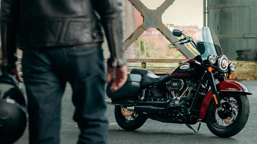 Ein Biker in Lederjacke hält einen Helm in der Hand und steht vor einer eleganten Harley-Davidson-Motorrad, das auf einer urbanen Straße parkt.