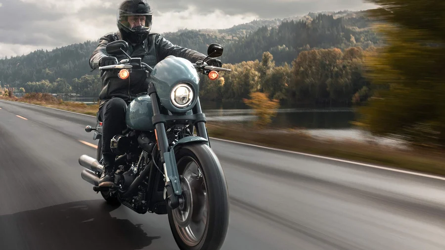Ein Motorradfahrer fährt mit einer Harley-Davidson auf einer malerischen Straße entlang eines Gewässers, umgeben von herbstlichen Bäumen.
