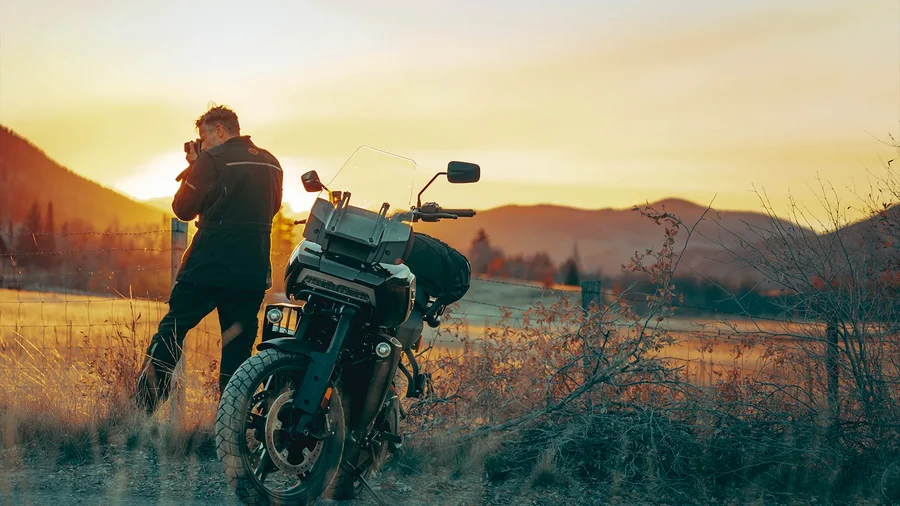 Ein Motorradfahrer steht mit dem Rücken zur Kamera und blickt bei Sonnenuntergang auf die Landschaft, während sein Motorrad neben ihm steht.
