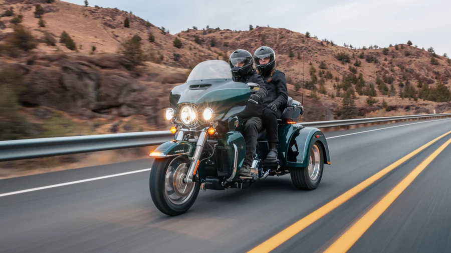 Ein grünes Harley-Davidson Motorrad mit Beiwagen fährt auf einer Landstraße, während zwei Fahrer Schutzhelme tragen und die Landschaft im Hintergrund sichtbar ist.
