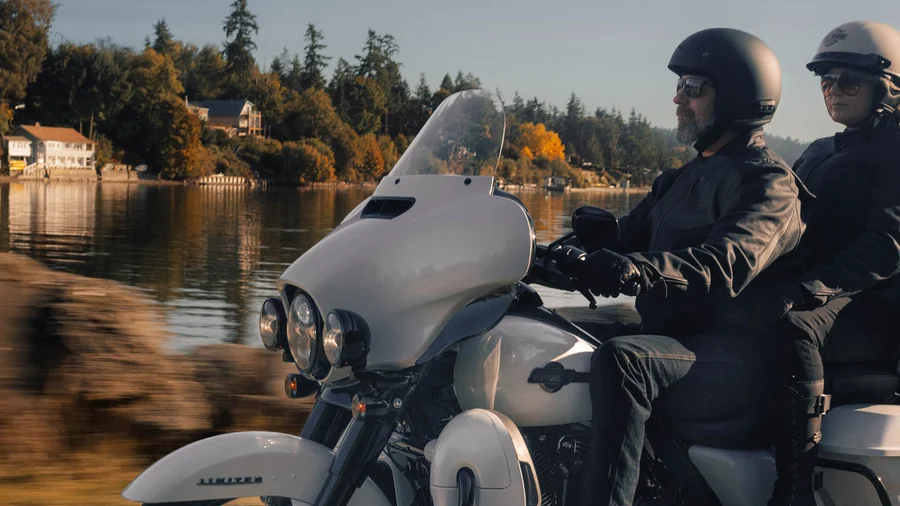Ein Paar fährt auf einem Harley-Davidson Motorrad entlang eines ruhigen Gewässers, umgeben von einer herbstlichen Landschaft.